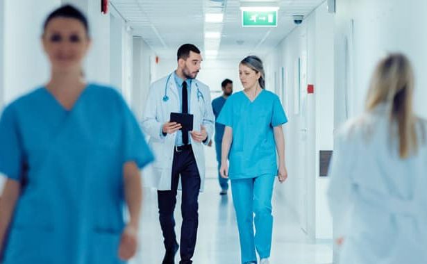 A doctor and nurse discussing patient data on a digital tablet in a modern hospital corridor, symbolizing Dedalus's high-performing EMR and clinical workflow solutions in the Australian healthcare market.